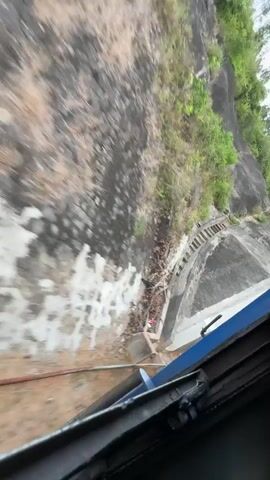 The video shows a view from inside a vehicle looking out at a steep, rocky, and overgrown hillside. A concrete structure with a series of steps is built into the side of the hill, running diagonally downwards. The vehicle appears to be moving, as evidenced by motion blur in the foliage and the concrete structure. The lower part of the frame shows the interior of the vehicle, including what appears to be a windshield wiper and part of the dashboard. The scene is predominantly green from the vegetation and gray from the rock and concrete.