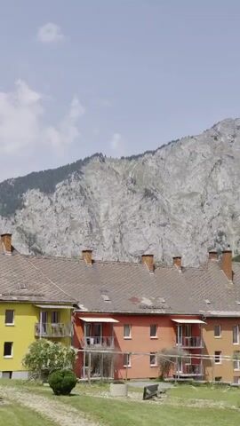 The video pans across a row of colorful apartment buildings with pitched roofs and chimneys. In the background, a large, rocky mountain covered with trees rises against a bright blue sky with scattered clouds. The buildings appear to be older, with some showing signs of wear. The area in front of the buildings is grassy with some trees and a few concrete circular structures. The scene depicts a residential area nestled in a mountainous landscape.