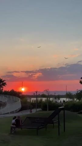 This is a vertical, candid shot of a scenic sunset over a body of water, likely a lake or bay, in Fethiye, Turkey. The sun, a vibrant orange orb, is low on the horizon, casting a fiery red and orange glow across the sky and clouds. Silhouetted mountains form a distant backdrop.

In the foreground, two people are sitting on a bench, seemingly enjoying the view. One person is wearing a headscarf, and they are positioned on the left side of the frame. Behind them, a contemporary-style curved walkway or bridge is visible, its white railing reflecting the warm light. To the right, a grassy slope leads down to a black metal bench and a park-like setting with trees and lampposts.

The time of day is clearly sunset, indicated by the low sun and the dramatic coloration of the sky. The weather appears clear and calm, ideal for outdoor enjoyment. Two birds are captured in flight against the colorful sky, adding a dynamic element to the scene. The overall mood is peaceful and serene, evoking a sense of relaxation and appreciation for nature's beauty. There is no visible text in the media.
