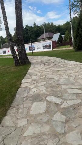 The video pans across a paved walkway made of irregular stones. To the left of the walkway is a well-maintained lawn dotted with trees. In the background, there is a white banner displaying various images, possibly advertisements or information panels. Beyond the banner, houses with reddish-brown roofs and dense green trees are visible, indicating a rural or suburban setting. The sky is partly cloudy. A few fallen leaves are scattered on the grass. The overall scene appears to be an outdoor park or recreational area.