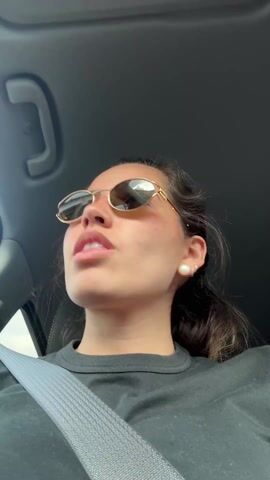 A young woman is seen from a low angle inside a car, wearing sunglasses and a black t-shirt. She is wearing a seatbelt. Her hair is pulled back. She is singing and looking upwards. The car's interior is gray. The video appears to be shot during the day.