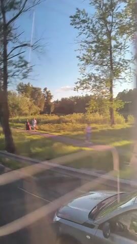 The video shows a view from a moving vehicle, likely a car, driving along a road. A guardrail is visible on the side of the road. Beyond the guardrail, there's a grassy field, with trees lining the edge. In the distance, a building with a spire or tower is visible, partially obscured by trees. The sun is shining brightly, creating lens flare and a general bright and sunny atmosphere.