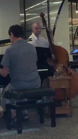 This media file captures a live musical performance, likely in Seville, Spain, as suggested by the context. The scene features two musicians, both men, engaged in playing instruments. In the foreground, a man with dark hair is seated at a piano, his back to the camera, playing the instrument. He is wearing a blue-grey short-sleeved shirt and light-colored trousers, with dark sandals on his feet. He is seated on a black piano bench.

Behind the pianist, another man, bald and wearing a white shirt, stands and plays a large upright bass. His focus is directed towards the instrument as he draws the bow across the strings. A small wooden side table is positioned next to the upright bass, holding several wine glasses.

The setting appears to be an indoor venue, possibly a restaurant or a lounge, with a dimly lit ambiance. Natural light streams in from large windows in the background, reflecting off surfaces and creating a slightly blurred effect. The architecture suggests a modern or renovated space, with exposed brick or concrete elements visible in the wall. The overall mood is one of focused musical performance, with the soft lighting and the presence of wine glasses hinting at a relaxed, evening setting. There is no visible text in the media file.