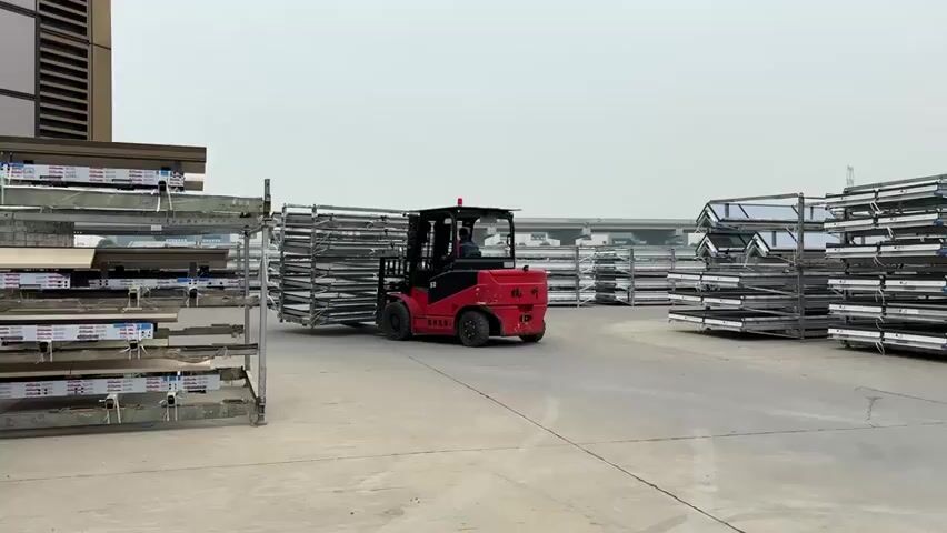 A red forklift moves in a gray concrete industrial area. The forklift is carrying a large stack of metal frames with multiple shelves. More stacks of these metal frames are organized around the area.  A tall building is visible in the background. An audible warning beep is present throughout the scene.