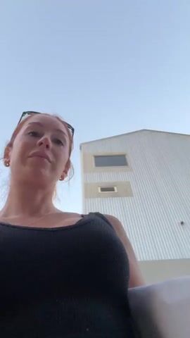 A woman with red hair and sunglasses on her head is filming herself with a smartphone. She is wearing a black tank top and appears to be outdoors. She is walking and the background shows a construction site or an area being set up for an event. There are white panels, ladders, and what looks like event signage in the background. She walks past a blue and white structure with signage that says "VISA tap in". In the final frames, she is walking past another person and says "Hi boyfriend".