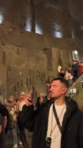 A group of people stand in a dimly lit stone room, potentially a cellar or ancient chamber. The walls are rough and textured, suggesting stonework. Several individuals are looking at the walls, some holding and using mobile phones, presumably to take photos or videos. The lighting is low, with some light appearing to be artificial. The camera moves, showing different members of the group.