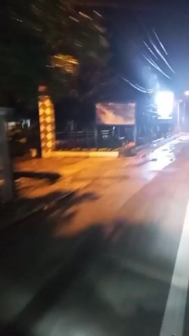 The video shows a nighttime scene from a moving vehicle. The road appears wet, reflecting the light from streetlights and a large, illuminated billboard. The billboard displays an image that is difficult to discern due to the low light and motion blur. Trees and utility poles are visible along the side of the road. The overall impression is of a drive through a dimly lit urban or suburban area at night.