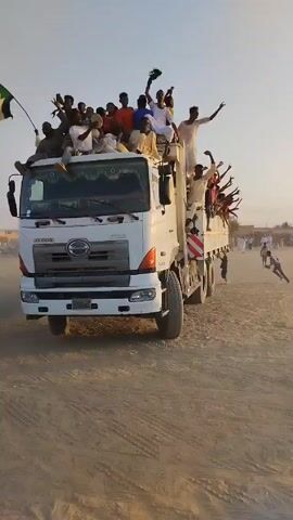 Here's a concise description of the video:

The video shows a white Hino truck driving across a dusty area. The truck is packed with people, many of whom are on the roof or in the truck bed, waving their arms and appearing to celebrate. Some of them are holding objects. There's a crowd of people visible in the background, and the sky is clear. The atmosphere seems lively and celebratory.