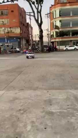The video pans up from a square showing a child driving a white electric toy car to a wide, low-angle shot of a tall, yellow and white church. The church has two tall towers with red domes on top. There are people walking around and sitting on the steps leading up to the church. The square in front of the church is paved with gray stones and has patches of green grass. The sky is blue.