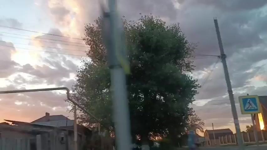 The video shows a view from inside a moving vehicle, looking out at a rural landscape. The sky is filled with dramatic clouds, illuminated by what appears to be sunset or sunrise. There are trees and houses visible, along with what looks like a soccer goalpost in the foreground. The footage is shaky, indicating movement, and the camera is angled slightly upwards.