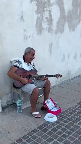 A man sits on a chair playing an acoustic guitar in front of a textured wall. He is wearing a white and black patterned shirt and grey shorts. In front of him, on the ground, is a pink case with "CD" written on it, a bottle of water, and some coins. The scene transitions to a narrow cobblestone street with a few people walking. Then, a group of people are seen sitting and standing by a stone wall overlooking the sea. Finally, the video shows a wire fence with a view of the ocean.