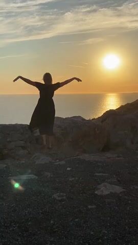 A person wearing a dark dress stands on a rocky cliff overlooking the ocean at sunset. The person has their arms raised in the air. The sun is setting over the water, casting a golden light. The sky is cloudy.