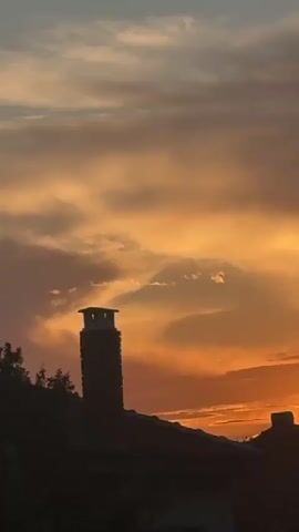 The video shows a sunset sky with orange and grey clouds. Silhouetted buildings, including a church steeple with a cross, are visible in the foreground. The camera pans upward and slightly rightward. Later shots show metal construction supports and telephone poles against the darkening sky.