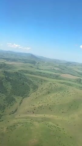 This video shows an aerial view of a vast, rolling landscape under a clear blue sky. The terrain is characterized by green, undulating hills and valleys. Patches of dense green foliage, likely trees, are scattered across the landscape, particularly in the mid-ground and left side of the frame. Further into the distance, the landscape appears more cultivated, with visible fields of varying shades of green and brown, suggesting agricultural activity. The horizon is marked by a chain of hazy, distant mountains. There are no people, vehicles, or man-made structures readily apparent in the visible portion of the video. The lighting suggests it is daytime, likely midday, given the bright sky and the way light falls on the landscape. The perspective is that of an elevated vantage point, as if from an aircraft or drone.