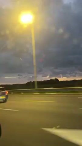 The video was shot from a moving vehicle on a highway. Other vehicles are visible on the road, mostly in the adjacent lane. Beyond the guardrail, a field stretches out to the horizon, topped by a dark line of trees. The sky is filled with dark clouds, but the setting sun peeks through, painting the horizon with streaks of orange and yellow. The video also picks up reflections of red objects from inside the vehicle. The lighting is dim and natural, with an added orange glow from the highway lights.