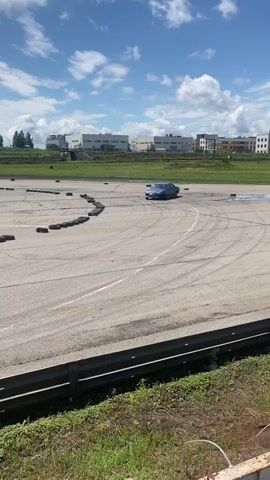 A purple sedan drifts on a paved track marked with tires. The car is in motion, kicking up smoke from its tires. The background shows industrial buildings, a cell tower, and a cloudy blue sky. The scene appears to be an outdoor racing or drifting event.