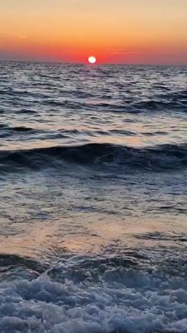 This is a vertical video of a sunset over the ocean, likely at a beach. The sun is a bright, round orb, appearing red and orange as it sits low on the horizon. The sky above transitions from a soft yellow near the sun to a pale orange and then a muted grey towards the top of the frame. The ocean is filled with choppy, dark blue waves, with some white foam visible as they crash onto the shore. The water reflects the warm colors of the sunset, creating streaks of orange and yellow on its surface. The foreground shows the wet sand and pebbles of the beach, covered in frothy white seafoam. The overall mood is serene and peaceful, evoking the beauty of nature at the end of the day. The location is identified as Zadar, Croatia. There is no visible text in the video.