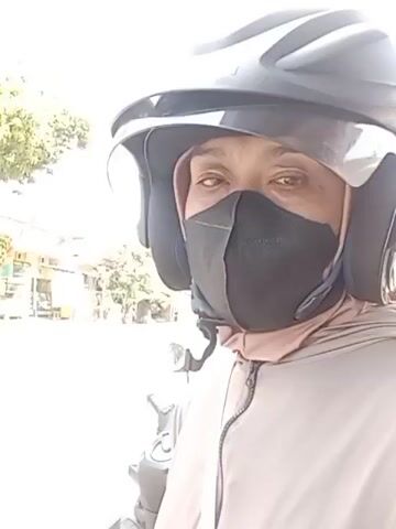 A person wearing a gray helmet and a black face mask looks into the camera. The person is also wearing a light brown hooded jacket. The background is blurry and appears to be an outdoor environment with some structures and trees. A portion of a motorcycle is visible in the lower left corner of the frame.