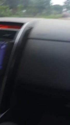 The video shows the interior of a car from the passenger's perspective. The dashboard features a touchscreen display, air vents, and various controls. A car accessory with a blue light is plugged into the car's power outlet. The driver's hands are visible on the steering wheel. The car appears to be in motion, with greenery visible through the windshield.