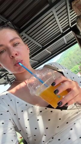 The video shows a woman drinking orange juice from a plastic cup with a blue straw. She is wearing a white dress with black polka dots. The woman has long brown hair and is wearing black nail polish. The setting appears to be outdoors, with a corrugated metal roof overhead and lush greenery in the background. The lighting suggests it is daytime, with sunlight casting shadows. The woman's expression is neutral as she sips her drink. The scene could be a cafe or restaurant with an outdoor seating area, possibly in a rural or scenic location. The background foliage suggests a warm climate. There is no visible text in the video.