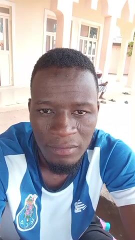 A man is taking a selfie outdoors. He is wearing a blue and white striped shirt. He has dark skin and short black hair. There are buildings and other people in the background. The man smiles and touches his forehead.