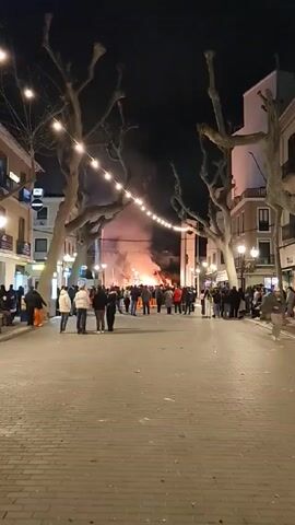 At night, a crowd of people stand around a large fire in the center of a brick-paved street. String lights hang over the street, lined by trees with bare branches. Buildings flank both sides of the street. Some people are sitting on the sides, while the majority are standing or walking toward the fire. Smoke from the fire rises into the night sky.