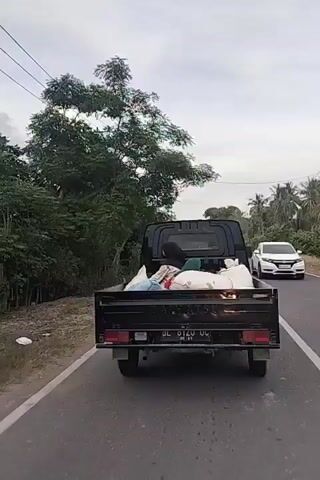 A black pickup truck is driving down a paved road. The truck is loaded with several large white sacks, some appearing to be filled with goods. A person wearing a green headscarf and a black jacket is sitting in the truck bed, partially obscured by the sacks. Trees and vegetation line the sides of the road, and utility poles are visible in the distance. The sky is overcast.
