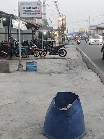 The video shows a street scene in what appears to be a town or city. In the foreground, a large, blue, damaged trash bin is visible, filled with debris. Further back, a concrete sidewalk runs alongside a road. On the sidewalk, several motorcycles are parked. A sign for "dr. ROLAS JAMES S. DOKTER UMUM" is prominently displayed above a small building, possibly a clinic or office. There are other stalls or shops along the sidewalk, some with lights on. A mosque, identified by its dome and minaret, is visible in the background. Traffic, including cars and motorcycles, moves along the road. The overall atmosphere suggests a relatively busy commercial area, possibly during the late afternoon or early evening, given the lighting conditions.