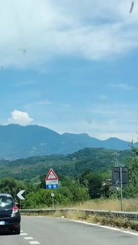 The video shows a view from inside a vehicle traveling on a road through a mountainous, green landscape under a bright blue sky with scattered clouds. In the foreground, the rear of a black Renault Clio is visible, moving along the right side of a paved road with white lane markings. A traffic sign with a yellow triangle and black arrows indicating a lane merge is visible on the right side of the road. Further down the road, other vehicles are traveling. Signs with black and white chevrons indicating sharp curves are visible along the roadside. A sign with a red triangle depicting slippery conditions and snowflakes is also visible on the right. In the background, rolling green hills and mountains rise, with some buildings visible in the distance. The overall scene suggests a scenic drive through a rural, possibly Mediterranean, environment.