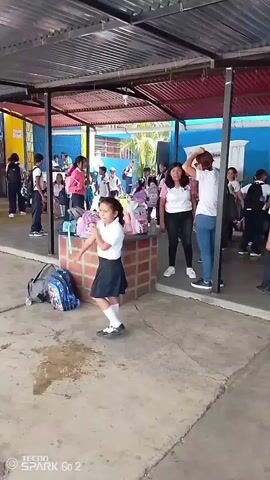 A group of students and a few adults are gathered in what appears to be an outdoor covered area of a school. Most of the students are wearing school uniforms, consisting of white shirts and dark-colored skirts or pants. Some students are wearing backpacks. Adults are mostly wearing casual clothes. Several backpacks and personal items are placed on a brick structure. A blue wall behind the group has the name of the school written on it, "Escuela Primaria Estatal Amatio Vivas, Salamanca." The environment looks like a schoolyard or a gathering place at a school. Some people are talking while others are moving around. The general activity level appears to be fairly relaxed and social.