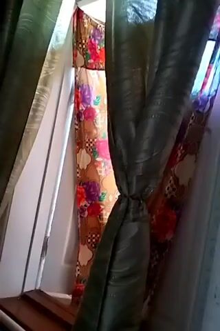 The video shows a room with curtains. A patterned dress is visible between two dark green curtains. The dress has a floral pattern with colors such as red, purple, and yellow. The dress is hanging on a rod, possibly in front of a doorway or window. The lighting suggests it might be daytime.