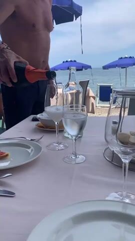 The video opens with a close-up view of a person's torso from the shoulders down. They are wearing dark pants and several bracelets on their left wrist. The person is holding a dark bottle with an orange cap and is pouring a bubbly white liquid into a clear wine glass. In the background, there is a table with a white tablecloth, plates, and glasses. One glass already contains a white, bubbly drink, and another contains clear liquid with ice. Further in the background, the ocean is visible, along with blue beach umbrellas and lounge chairs. The shot then pans slightly to the right, showing more of the table setting and the beach scene. The person continues to pour the drink, and the bottle is lifted. The camera then continues to pan right, focusing on the table and the ocean view, with several empty wine glasses and bread on a plate. The video ends with a wider shot of the table and the beach.