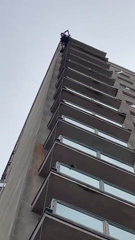 The video shows a tall, concrete apartment building with numerous balconies. A person is visible near the top of the building, secured by ropes, possibly performing maintenance or repairs. The camera is angled upwards, emphasizing the height of the building. The sky is overcast.