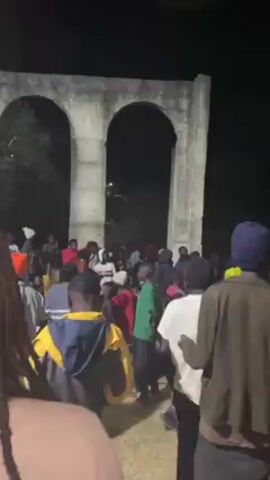 The video depicts a large group of people gathered outdoors at night. They appear to be congregated around a structure that resembles an archway. The lighting is dim, making it difficult to discern specific details about individuals' clothing and features. There is some movement within the crowd, suggesting interaction or activity, but the exact nature of this activity is not clear from the video. The scene is shot from within the crowd, offering a ground-level perspective of the gathering.