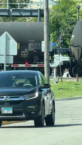 The video shows a train crossing a road. The camera is inside a car stopped at the intersection. The first rail car is a large black tank car labeled “SR 13450”. The railroad crossing lights are flashing red. A person is visible in the car's side view mirror, wearing a blue shirt. The next rail car is a red boxcar labeled “83183”. The next tank car is labeled “GATX 96029”. A person is briefly visible walking across the tracks as the train passes.