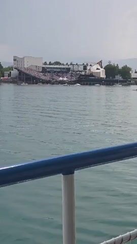 The video shows a wide view of a body of water with a large outdoor seating area on the shore. A crowd of people are seated in the tiered stands. Several boats are visible on the water in front of the seating area. The sky is overcast.