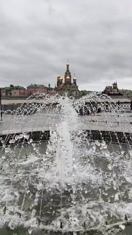 A vertical shot shows a fountain in the foreground spraying water upwards, with a cityscape in the background. The cityscape features buildings with domes, including a prominent golden-domed church, and appears to be on the bank of a body of water. The sky is overcast. The water in the fountain is translucent and shows individual droplets and streams.