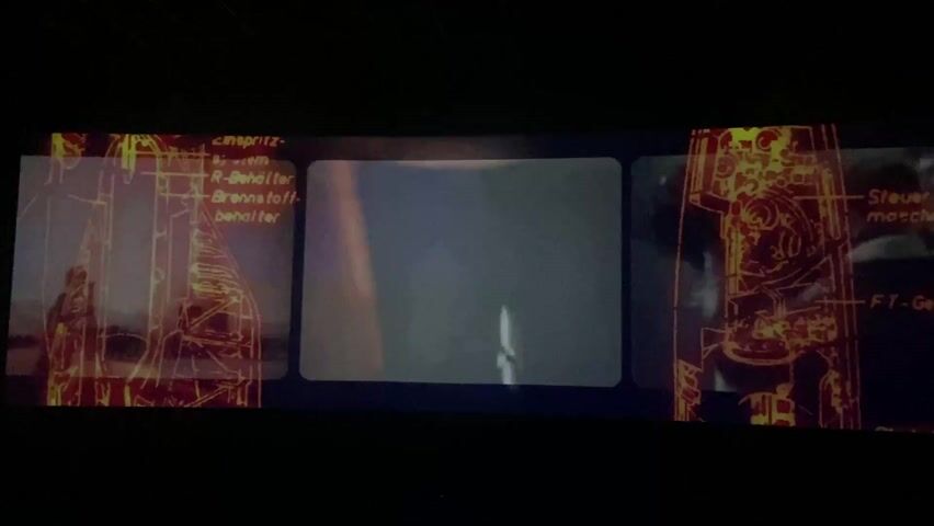 The video shows a screen in a dark room. First, a vintage newspaper is displayed with the headline "SPACE MAN ENTERS SPACE." The headline is surrounded by pictures of early astronauts. Then, the video shows a man in a white coat working on a space rocket in a hangar.