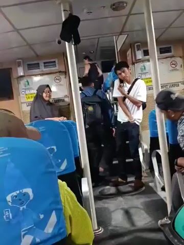 The video shows the interior of what appears to be a ferry or bus. Several people are visible, including a young man in a white t-shirt and jeans, talking on a cell phone and holding a bag of food. A woman in a hijab is seated on the left, and other passengers are seen on stairs and in seats. The seating is blue and has cartoonish images on the headrests. There are signs on the walls, ventilation grilles, and a television screen mounted high on the wall. The overall lighting is artificial and somewhat dim. The camera angle is at eye level, looking towards the stairs and the passengers.