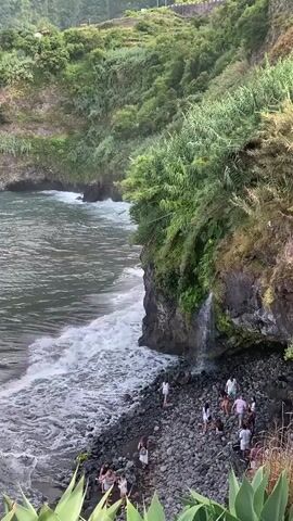 A high-angle, medium shot shows a rocky beach adjacent to a lush, green cliff face where a small waterfall cascades down. The ocean water is choppy and grey, with white waves crashing against the shore. A group of approximately ten people, mostly wearing light-colored clothing, are scattered on the dark, pebble-strewn beach. Some appear to be standing near the base of the cliff, while others are closer to the water's edge. The overall impression is of a natural, somewhat wild coastal environment with a few individuals enjoying the scenery.
