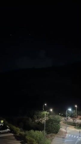 The video shows a nighttime outdoor scene. In the foreground, there is a parking lot with a white car parked on the left. In the middle ground, there are trees and bushes illuminated by streetlights. Two streetlights are clearly visible, one taller and one shorter, casting a warm glow. A paved road with white lines is visible to the right. In the background, there is a dark, silhouetted hill or mountain. Above the hill, the night sky is visible, with two faint green lights, possibly stars or distant aircraft, visible at the top of the frame. Some wispy clouds are present in the dark sky. The overall impression is a quiet, dimly lit night scene in a semi-rural or suburban area.