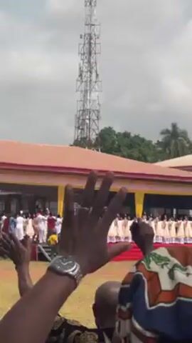 The video shows a large choir singing in front of a building with a red roof and yellow curtains. The choir members are wearing white and gold robes. People are standing on a red carpet in front of the stage. A person in the foreground is filming the scene with their hands raised. In the background, there is a tall communications tower. The environment appears to be outdoors on a grassy lawn.