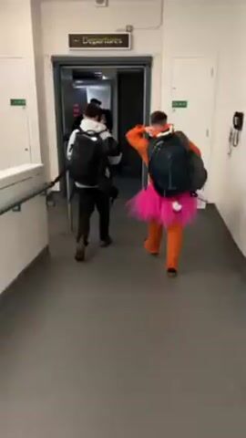 A group of people walk through a "Departures" doorway in a building, likely an airport. The person in the foreground is wearing an orange fox costume with a pink tutu and a white tail attached. They are carrying a dark backpack. Other people are ahead of them, also with backpacks. The walls are white, and the floor is gray. A sign labeled "LB 19 3" is visible on a doorframe to the right. The doorway has a metal frame and a black barrier post is visible in the left foreground.