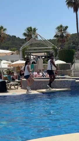 The video shows a daytime scene at a resort swimming pool. Two instructors, a woman and a man, are standing on a yellow mat at the edge of the pool, demonstrating exercises. They are wearing athletic clothing. The woman is wearing a white tank top and dark shorts, and the man is wearing a white tank top and dark shorts. Both are wearing white sneakers. Behind them, several people are relaxing on lounge chairs under umbrellas. In the pool, a group of people, including adults and children, are participating in an aqua aerobics class. They are moving their arms and legs in unison. The pool is surrounded by lush greenery, including palm trees and hills in the background. The sky is clear and blue, indicating sunny weather. The overall atmosphere appears to be lively and recreational.