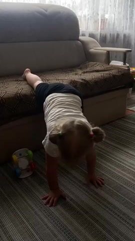 A young child is on all fours in front of a sofa, with their upper body tucked underneath it. The child has blonde hair in pigtails and is wearing a white t-shirt and black shorts. The child has an orange pacifier in their mouth. The child is on a striped carpet, and to the left of the child is a colorful toy. The sofa is upholstered in a brown and beige patterned fabric. The background shows a window with sheer curtains and a wall with a small table. The scene is lit by natural light.