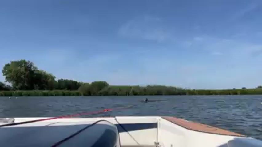 The video shows a person water skiing behind a boat on a sunny day. The boat is moving forward, pulling the skier through the water, creating a spray behind them. The sky is blue with a few scattered clouds.  Trees and reeds line the edge of the waterway.