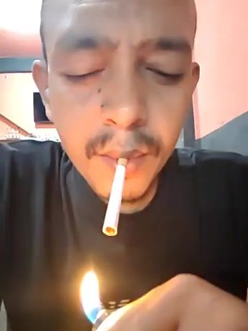 A medium close-up shot depicts a man lighting and smoking a cigarette. The man is fair-skinned, has a shaved head, and a mustache. He wears a black t-shirt. He lights the cigarette with a blue flame lighter. He takes a drag of the cigarette, expels the smoke, and then removes the cigarette from his mouth. The background features a wall that is part gray and part orange. A television screen and glassware are also visible in the background.