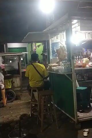 The video shows a nighttime scene at what appears to be an outdoor food stall or small shop. A person in a yellow shirt is sitting on a stool facing away from the camera, towards the stall. The stall has a counter with various items displayed, including what looks like bottled water and packaged goods. There are lights illuminating the stall. In the background, another person wearing a hooded jacket is visible behind the counter, possibly working or interacting with customers. The environment appears to be a street or alleyway with some debris on the ground. The video is dimly lit, and the camera is positioned at an angle.