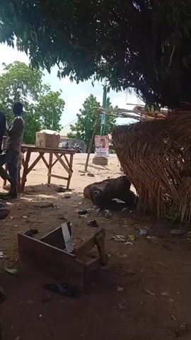 The video shows an outdoor scene with several people standing around a makeshift wooden table. The ground is dusty and littered with debris, including a broken wooden box. In the background, there are trees and a poster on a pole. The lighting suggests it is daytime. Two men are standing at the table, and another person is to their left. The man on the right of the table appears to be holding something. There is also a thatched structure to the right of the frame.