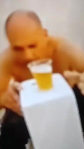 A blurry video shows a bald man, shirtless, leaning forward. He is holding a clear plastic cup filled with a yellow liquid, possibly beer, which is placed on top of a white rectangular object. In his right hand, he is holding another clear object that is being poured into the cup on the white object. The man's face is in focus, but his body is out of focus. The background is also blurry and appears to be an outdoor setting.